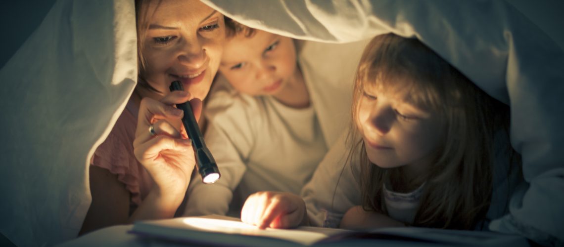 A woman and two children are reading in bed under a sheet.  The woman is holding a small black flashlight.  The little boy is near the mother.  Next to him, the little girl is holding her finger on the page of the book.